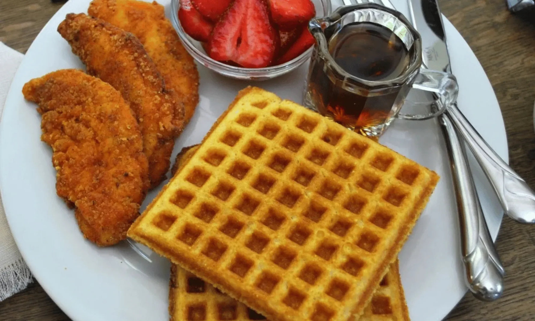 Gluten-free cornbread waffles on a white plate with fried chicken tenders, fresh strawberries, and a pitcher of maple syrup