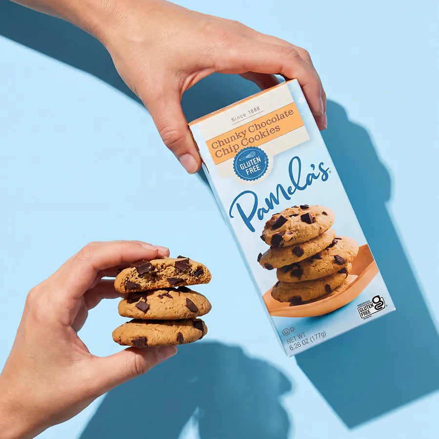 A hand holds a box of Pamela's Chunky Chocolate Chip Cookies against a light blue background, while another hand holds a small stack of chocolate chip cookies in the foreground.