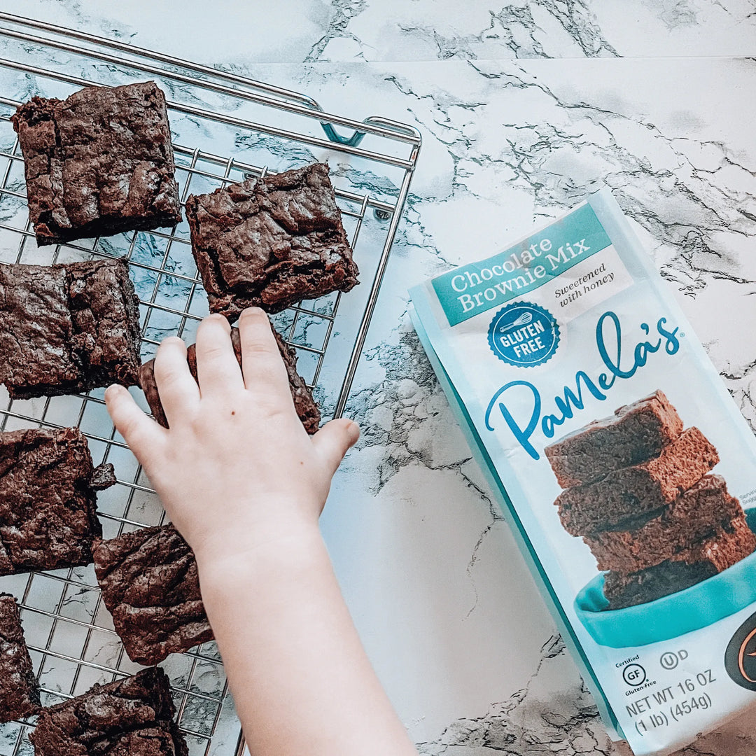 Chocolate brownies on a cooling rack with a box of Pamela's Chocolate Brownie Mix on a marble surface.
