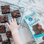 Chocolate brownies on a cooling rack with a box of Pamela's Chocolate Brownie Mix on a marble surface.