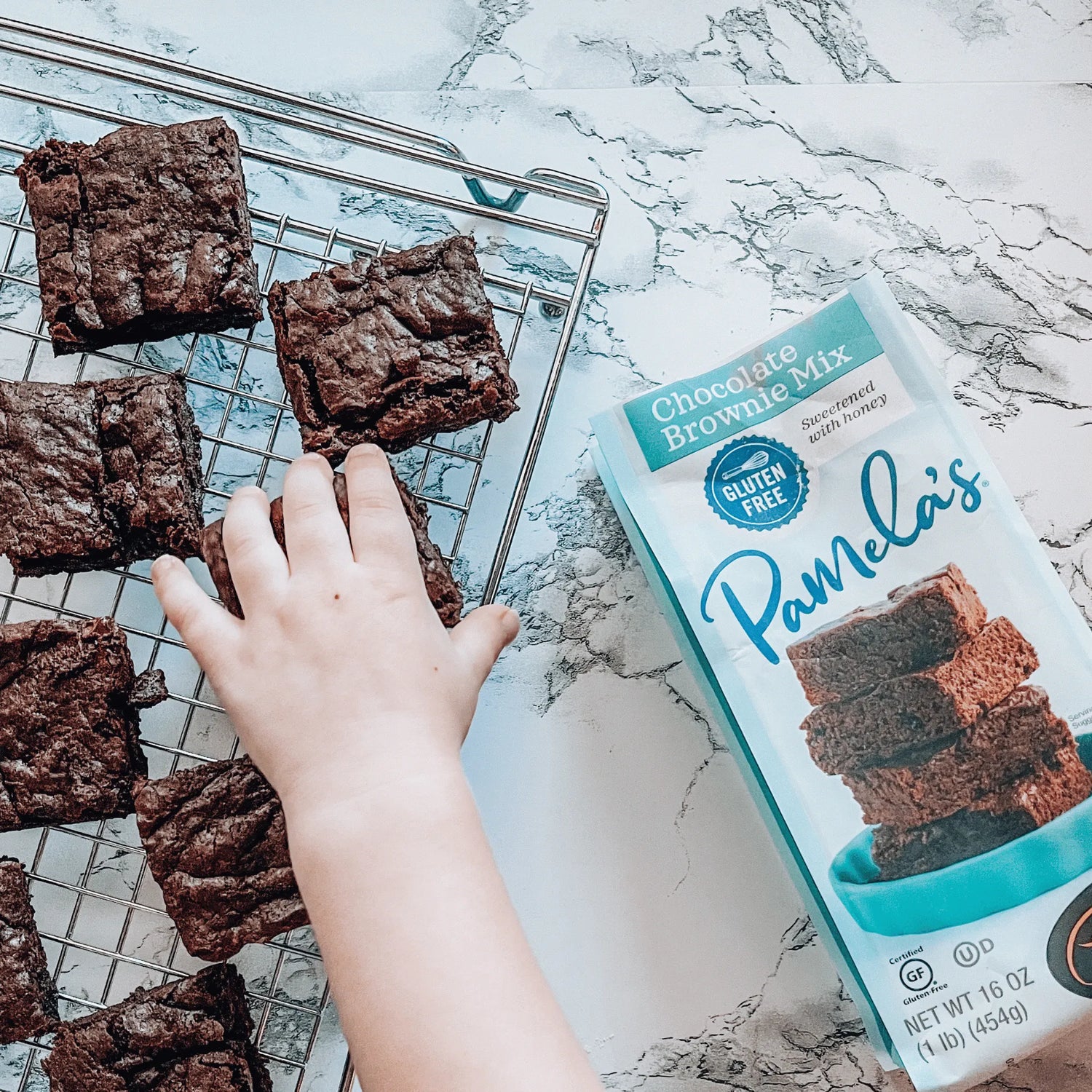 Chocolate brownies on a cooling rack with a box of Pamela's Chocolate Brownie Mix on a marble surface.