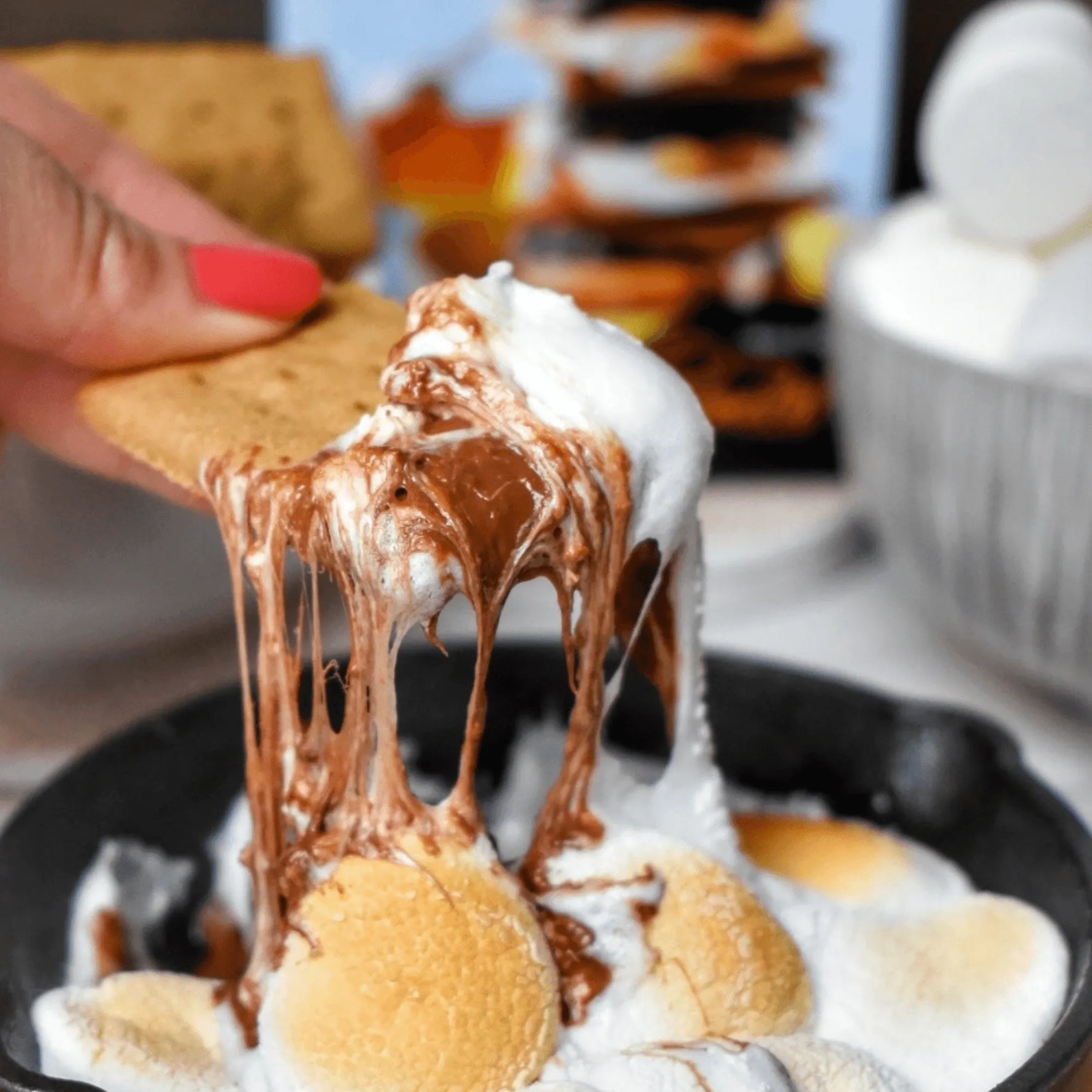 Hand dipping a graham cracker into melted chocolate with marshmallows on a plate.