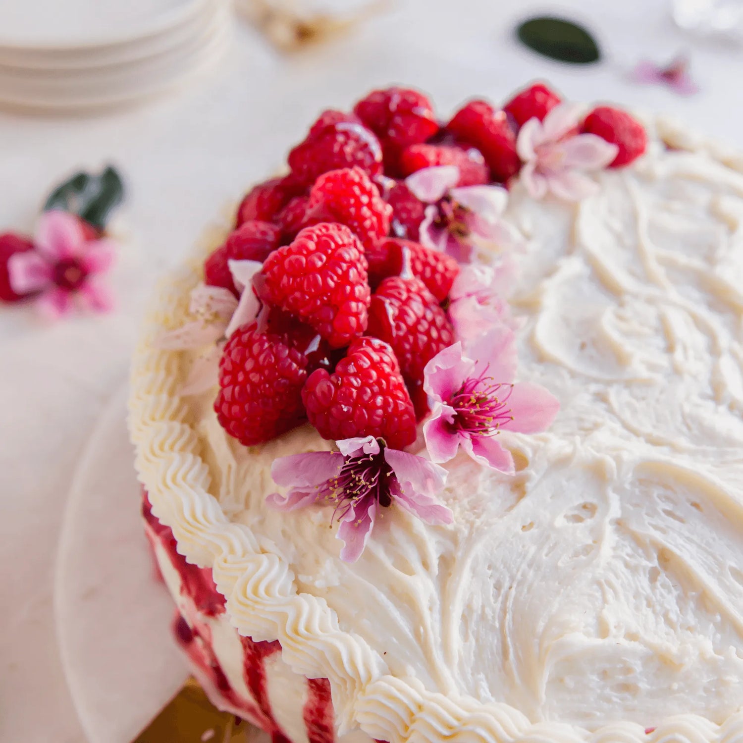 Decorative cake with white frosting, raspberries, and pink flowers on a light background.