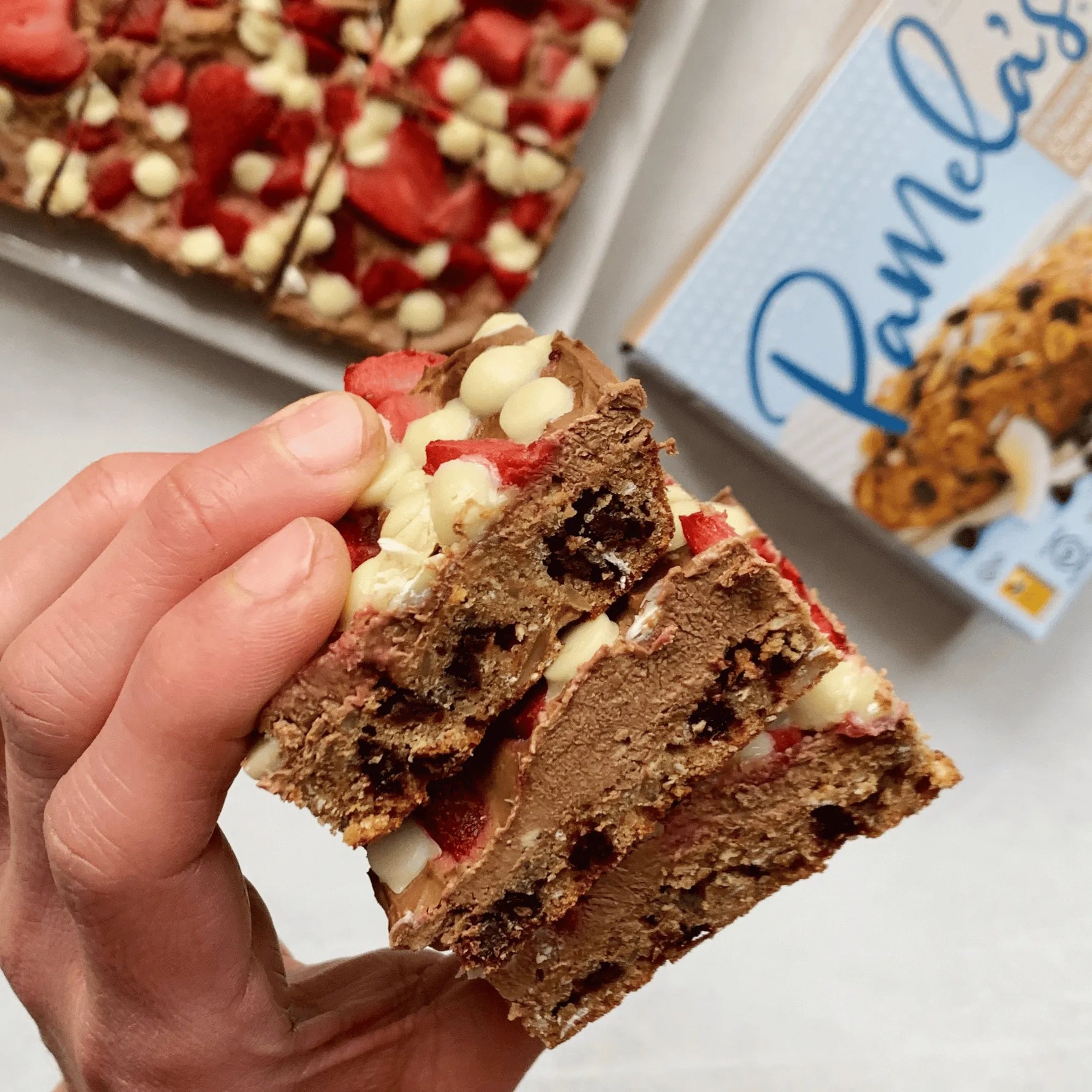 Hand holding a chocolate cookie bar with strawberries and white chocolate chips, with a box of Pamela's products in the background.