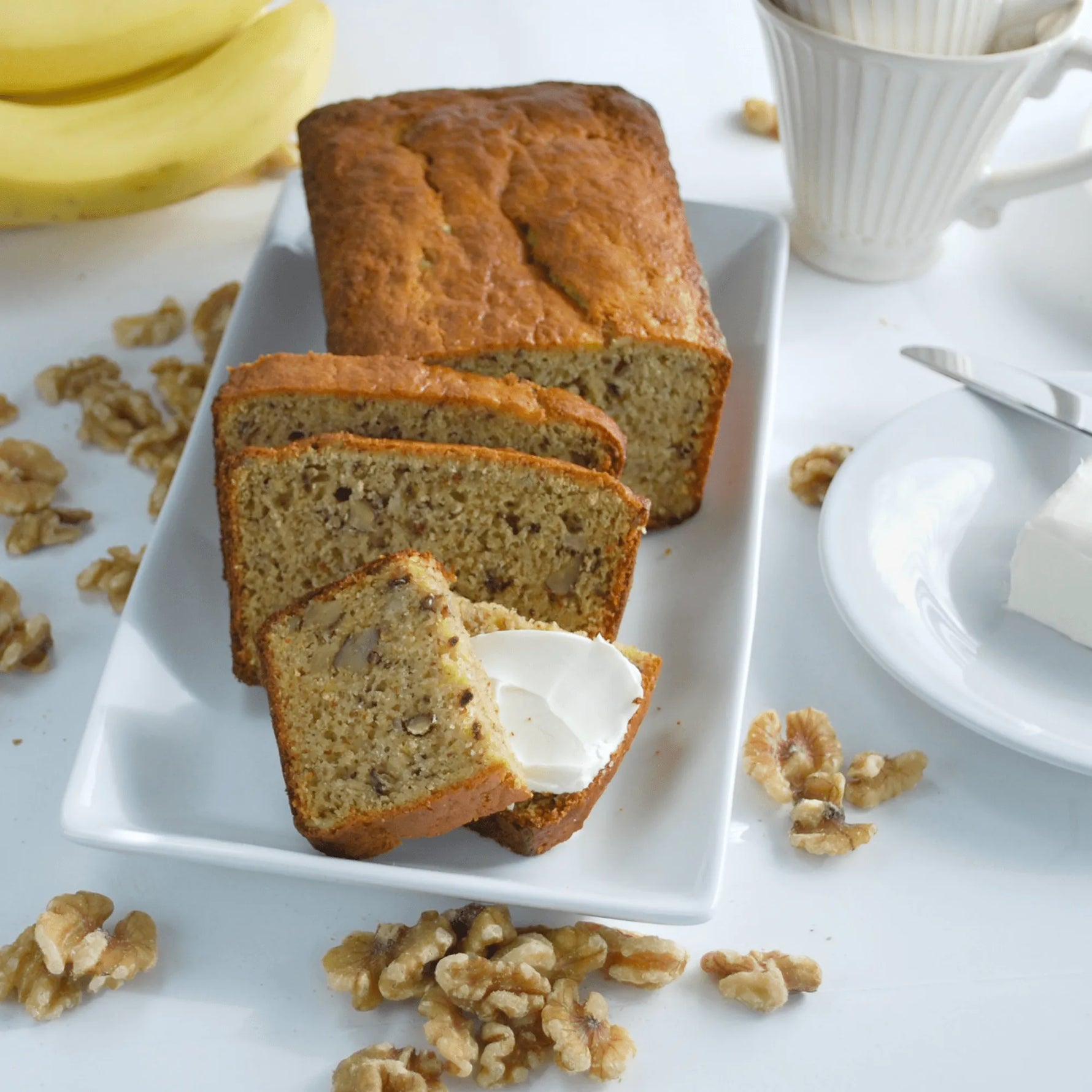 Loaf of banana bread with slices on a plate, surrounded by bananas and walnuts on a white surface.