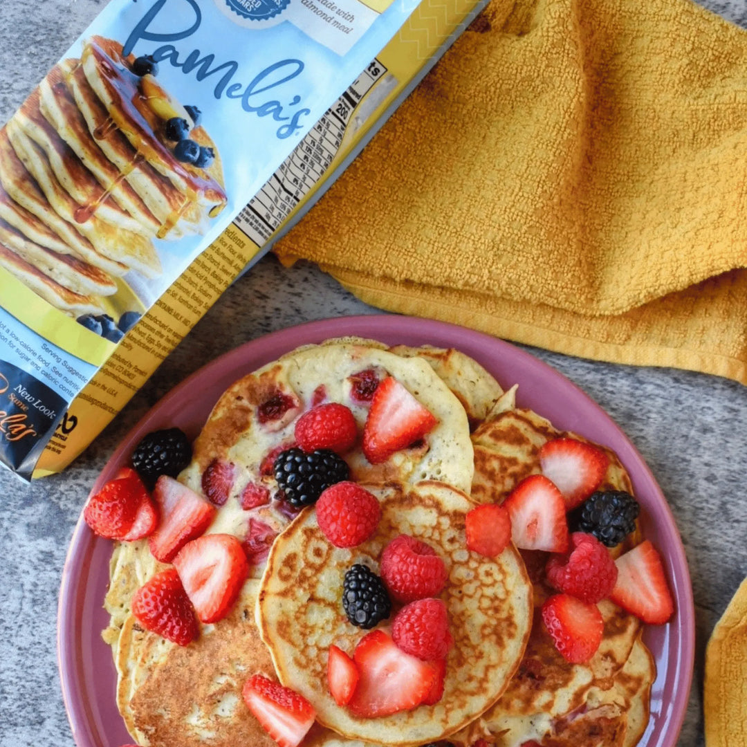 Stack of pancakes with berries on a pink plate next to Pamela's Pancake Mix packaging.